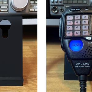 Puede incluir: Soporte negro con un recorte en forma de ojo de cerradura, que sostiene un transceptor FM de banda dual negro. El transceptor tiene un teclado, un botón azul iluminado y el texto "DUAL BAND FM TRANSCEIVER".