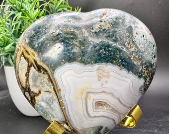 May include: A heart-shaped stone sculpture with a polished surface, featuring a mix of green, white, and brown colors. The stone is displayed on a gold-colored metal stand. The sculpture is a decorative item.