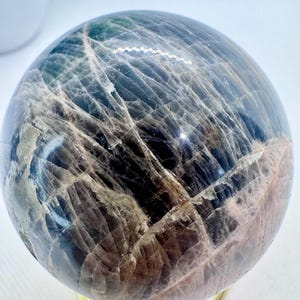 May include: A large, polished, gray and white moonstone sphere on a gold metal stand. The sphere has a unique, layered pattern and a reflective surface.