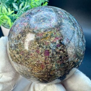 May include: A polished sphere of ruby in matrix, a type of gemstone. The sphere is a deep green with red ruby inclusions and a speckled gold pattern.