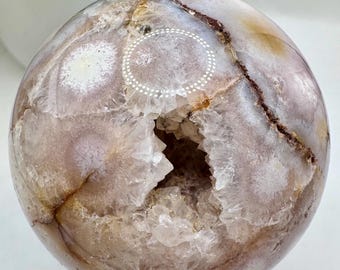 Puede incluir: Una gran esfera de geoda de ágata marrón y blanca con una abertura natural que revela un interior forrado de cristales. La esfera está pulida y se asienta sobre un soporte negro.