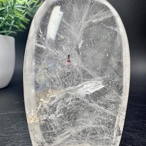 May include: A clear quartz crystal, rounded at the top and flat at the base. It features internal fractures and inclusions, with a small pink mineral visible. The crystal is set against a dark backdrop.
