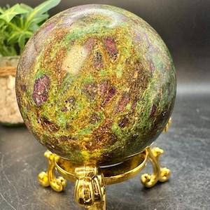 May include: A large, polished gemstone sphere with a mottled pattern of green, gold, and deep red. The sphere is displayed on a decorative gold-colored metal stand with ornate, curved legs. The overall aesthetic is one of natural beauty and luxury.