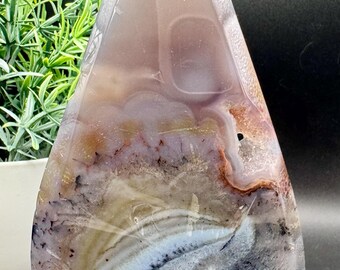 May include: A teardrop-shaped agate stone with a polished surface. The stone features layers of color, including shades of gray, white, brown, and purple. The top of the stone is clear, and the bottom is rounded. A decorative item.