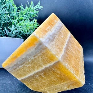 May include: A large, faceted, golden calcite crystal with bands of white and tan. The crystal is cut into a geometric shape, showcasing its natural layers and textures. The background is dark, highlighting the crystal's warm tones.