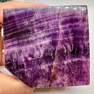 May include: A purple fluorite crystal with white and gold veins. The crystal is cut into a square shape with rounded edges.