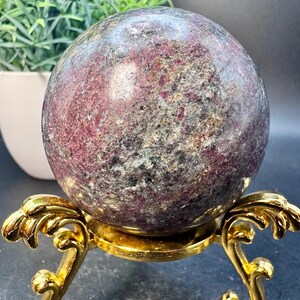 May include: A large, dark purple and gray gemstone sphere on a gold metal stand. The sphere has a speckled appearance with a mix of dark and light purple and gray colors.