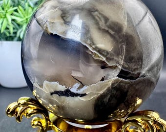 Peut inclure: Une sphère polie avec des motifs tourbillonnants de gris, de noir et de crème. L'objet est présenté sur un support décoratif en métal doré avec des détails courbes et ornés. Un petit pot de plante est en arrière-plan.