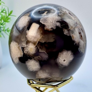 May include: A polished agate sphere with a dark gray base and white and light purple agate inclusions. The sphere is displayed on a gold metal stand.