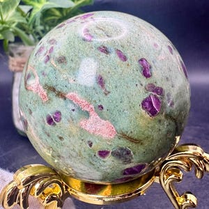 May include: A green sphere with deep purple and pink inclusions, resting on a gold-colored decorative stand. The sphere's surface has a mottled appearance, with varying shades of green and pink. The stand has an ornate, scrolled design.