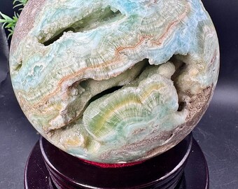 May include: A spherical, polished stone with a geode-like appearance. The orb features layers of blue, green, brown, and white hues. The stone is displayed on a dark wooden stand, showcasing its natural beauty and intricate patterns.