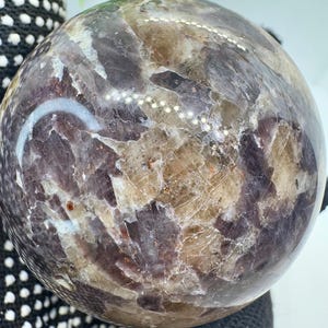 Puede incluir: Una esfera pulida con un patrón de mosaico en tonos marrones, beige y morados. La superficie tiene un aspecto natural y texturizado. Esta esfera decorativa podría ser un pisapapeles o una pieza de exhibición, de unos 10 cm de diámetro.