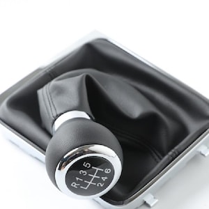 Może przedstawiać: Czarny skórzany kołnierz dźwigni zmiany biegów z chromowaną obwódką i srebrną gałką zmiany biegów z nadrukowanymi numerami R, 1, 3, 5 oraz 2, 4, 6.