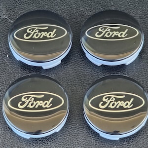 Könnte beinhalten: Vier schwarze Radkappen mit einem goldenen Ford-Logo. Die Radkappen haben eine glatte, glänzende Oberfläche.