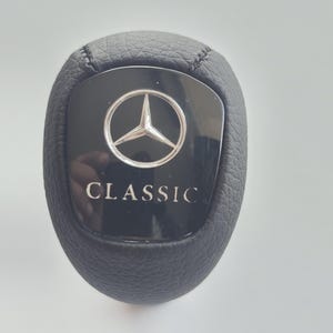 Könnte beinhalten: Schwarzer Leder-Schalthebelknauf mit silbernem Mercedes-Benz-Logo und dem Schriftzug "CLASSIC" in weiß.