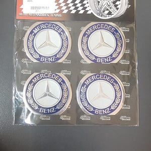 May include: Four silver Mercedes-Benz car emblems with blue trim and the words "Mercedes-Benz" in white lettering. The emblems are packaged in a clear plastic bag with a black and white checkered background.