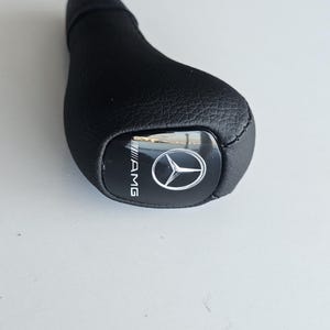 Op de afbeelding: Zwarte leren versnellingspookknop met een zilveren AMG-logo.