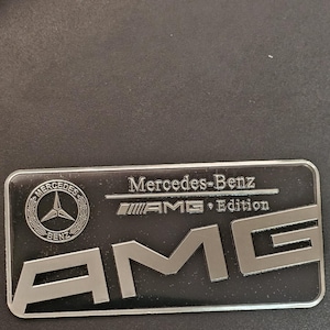 Könnte beinhalten: Ein silbernes Mercedes-Benz AMG Edition Emblem mit schwarzem Hintergrund. Das Emblem zeigt das Mercedes-Benz Logo und den Text "AMG Edition".