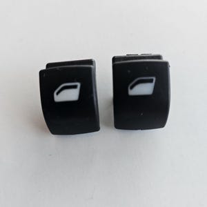 May include: Two black plastic car window switches with a white window icon on each.