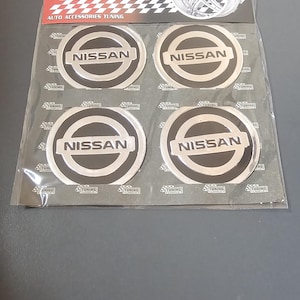 Könnte beinhalten: Eine Packung mit vier silbernen Nissan-Autoemblemen aus Metall. Die Embleme sind rund und haben einen schwarzen Hintergrund mit einem silbernen Nissan-Logo. Die Packung ist mit "Metal Series Auto Accessories Tuning" beschriftet.