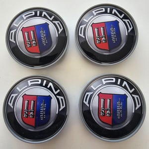 Puede incluir: Conjunto de cuatro tapacubos de coche negros y plateados con un logotipo rojo, blanco y azul que dice "ALPINE" en un círculo.
