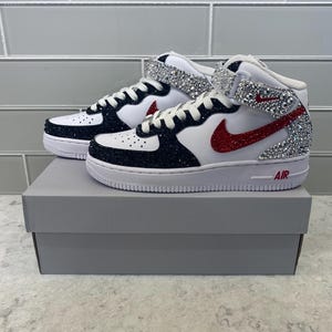 May include: A pair of white and black mid-top sneakers with red accents. The sneakers feature a sparkling silver strap and a red Nike swoosh. The word "AIR" is printed in red on the white sole.