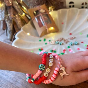 May include: A set of four colorful bracelets with red, pink, white, and green beads. One bracelet has the word "HO HO" spelled out in gold letters. Another bracelet has a peppermint candy design. The bracelets are stacked on a person's wrist with a gold reindeer charm.