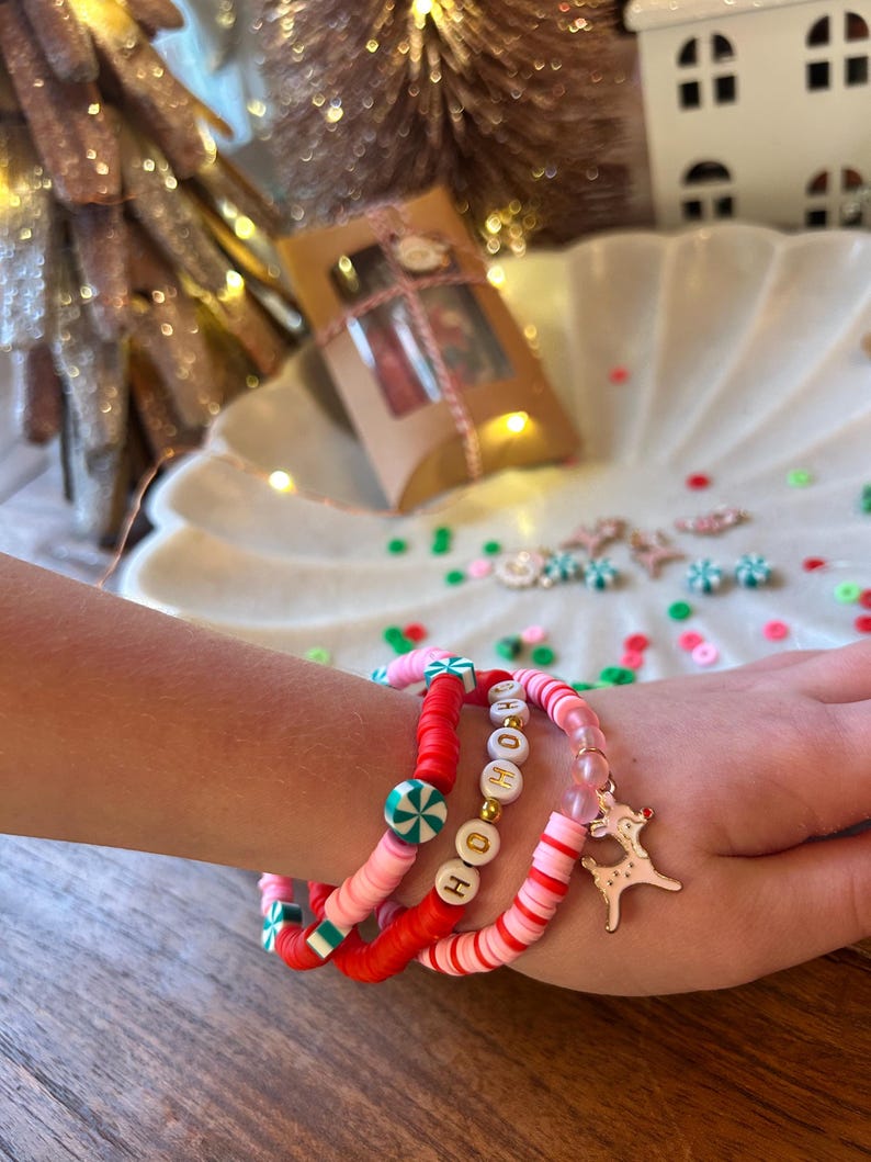 May include: A set of four colorful bracelets with red, pink, white, and green beads. One bracelet has the word "HO HO" spelled out in gold letters. Another bracelet has a peppermint candy design. The bracelets are stacked on a person's wrist with a gold reindeer charm.