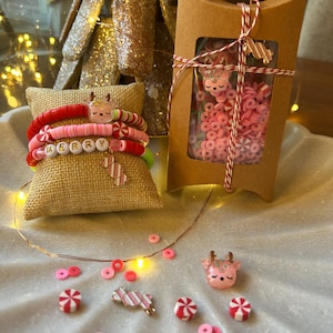 Puede incluir: Un conjunto de pulseras con tem&aacute;tica navide&ntilde;a con cuentas rosas, rojas y blancas, incluyendo un dije de bast&oacute;n de caramelo y un dije de reno. Las pulseras se muestran en una caja cubierta de arpillera con la palabra "MERRY" escrita en letras doradas. El conjunto tambi&eacute;n incluye un dije de reno rosa, dijes de bast&oacute;n de caramelo y cuentas rosas y blancas.