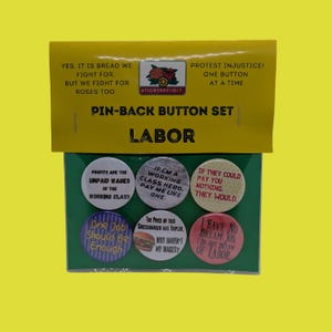 May include: A set of six round pin-back buttons on a yellow and green display card labeled Pin-Back Button Set Labor. The buttons feature various slogans about labor and wages, including text like Profits are the unpaid wages of the working class.