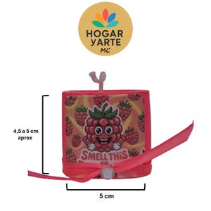 Puede incluir: Un pequeño producto cuadrado rosa con un gráfico de frambuesa y el texto "SMELL THIS". El producto mide aproximadamente 5 cm de ancho y 4,5-5 cm de alto. Se adjunta una cinta rosa. El producto tiene un pequeño asa rosa claro.