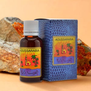 May include: A small brown glass bottle with a white cap, labelled "Aoussarabia" in Arabic and English. The bottle is sitting on a rock next to a blue box with the same label. The box has a textured surface and a scene of a mosque and palm trees.