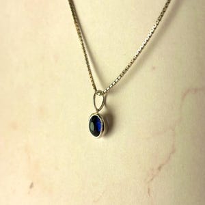 May include: A silver-toned box chain necklace featuring a small, round, bezel-set pendant with a deep blue gemstone. The pendant hangs from a simple silver-toned bail attached to the delicate, square-linked chain, creating a clean, simple look.