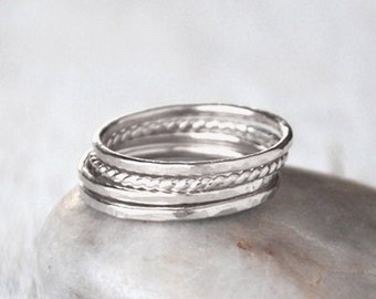 May include: A stack of silver rings with various textures, including smooth and twisted designs, rests on a smooth, gray stone. The rings are displayed against a soft, blurred background, creating a minimalist aesthetic. The rings are likely made of sterling silver.