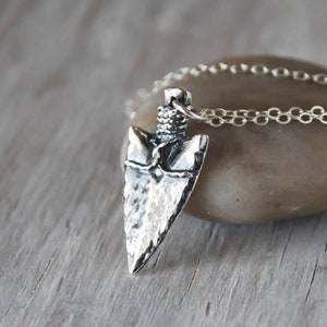 May include: Silver necklace with a hammered arrowhead pendant. The necklace is on a gray stone surface.