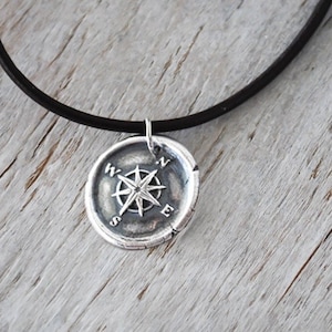 May include: A silver compass pendant necklace with a black leather cord. The compass features a star-shaped design with the letters N, S, E, and W around the edge.