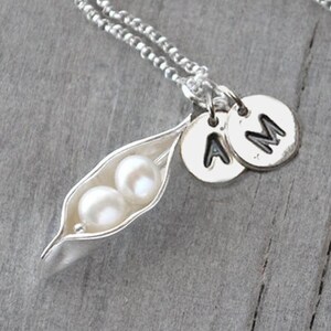 May include: A silver necklace with a pea pod charm containing two white pearls. The necklace also has two silver charms with the letters "A" and "M" on them.