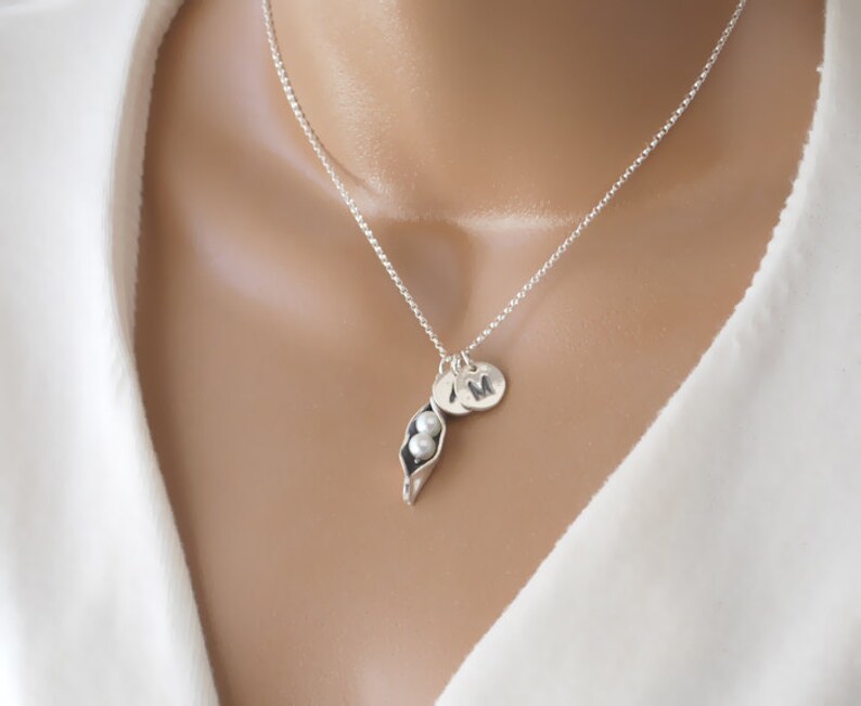 May include: A silver necklace with a pea pod charm and two pearl beads. The necklace also has a silver initial charm with the letter "M" on it.