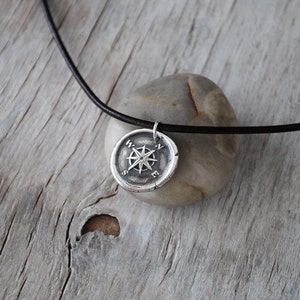 May include: A silver compass pendant necklace with a black leather cord. The compass features a star design with the letters N, S, E, and W around the edge.