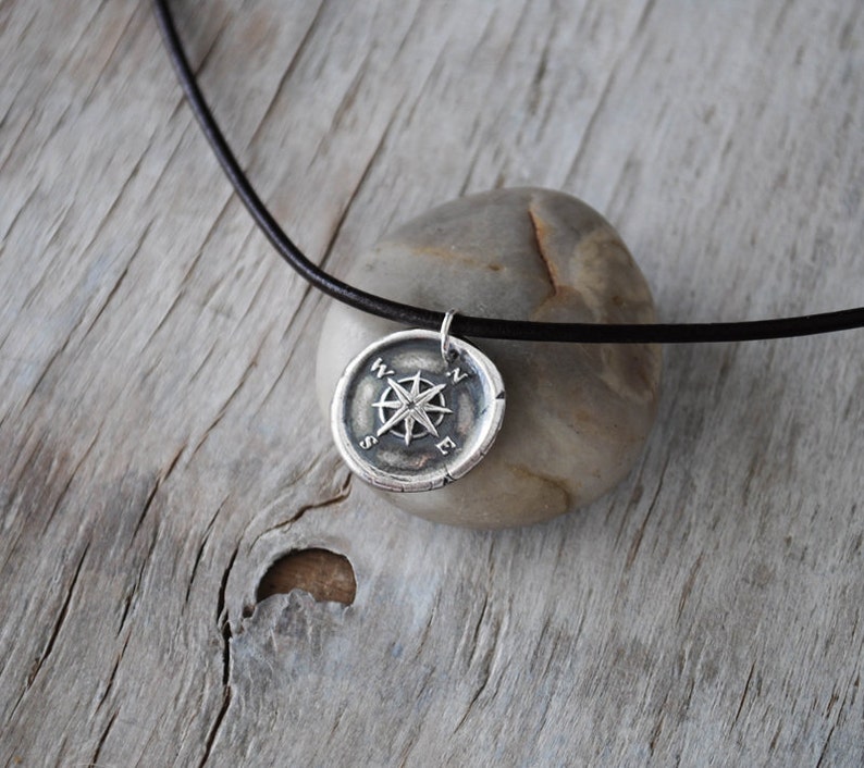 May include: A silver compass pendant necklace with a black leather cord. The compass features a star design with the letters N, S, E, and W around the edge.