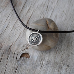 May include: A silver compass pendant necklace with a black leather cord. The compass features a star design with the letters N, S, E, and W around the edge.