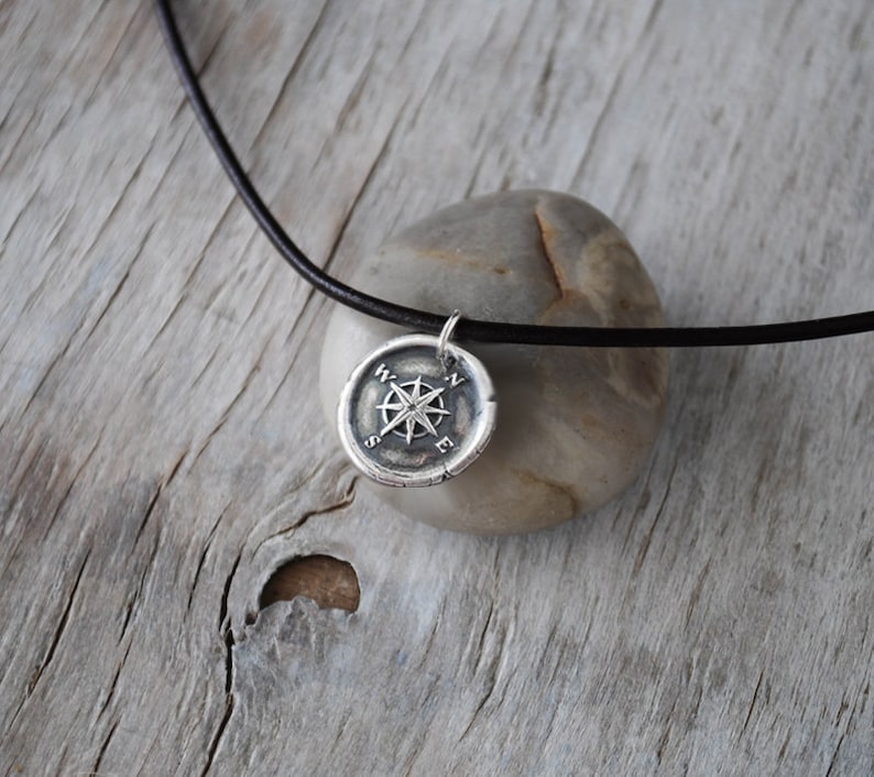 May include: A silver compass pendant necklace with a black leather cord. The compass features a star design with the letters N, S, E, and W around the edge.