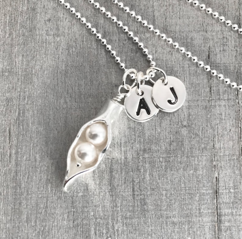 May include: A silver necklace with a pea pod charm and two pearl beads. The necklace has two initial charms, one with the letter 'A' and the other with the letter 'J'.