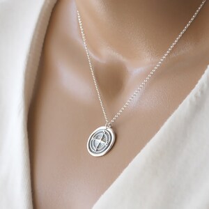 Puede incluir: Un collar de plata con un colgante redondo que presenta un dise&ntilde;o de br&uacute;jula. El colgante tiene una superficie texturizada y una cadena de plata.