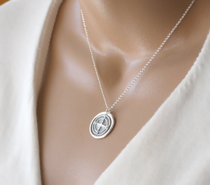 Puede incluir: Un collar de plata con un colgante redondo que presenta un dise&ntilde;o de br&uacute;jula. El colgante tiene una superficie texturizada y una cadena de plata.