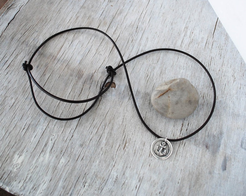 May include: A black leather cord necklace with a silver anchor pendant. The pendant is round and features a raised anchor design.
