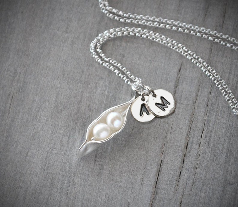 May include: A silver necklace with a pea pod charm containing two white pearls. The necklace also has two silver charms with the letters "A" and "M" on them.