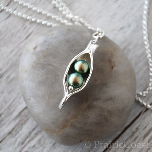 May include: A silver necklace with a pea pod charm. The charm has two green iridescent pearls inside. The necklace is on a white wooden surface.