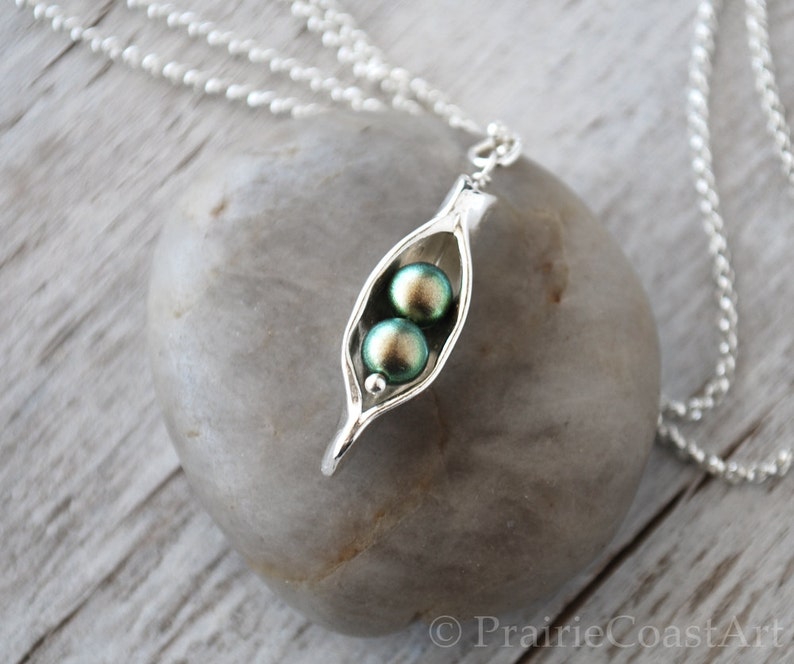 May include: A silver necklace with a pea pod charm. The charm has two green iridescent pearls inside. The necklace is on a white wooden surface.