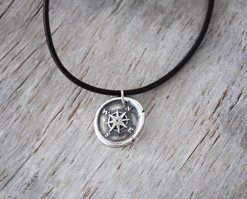 May include: A silver compass pendant necklace with a black leather cord. The compass features a star-shaped design with the letters N, S, E, and W around the edge.